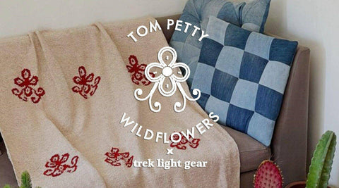 A tan blanket with red wildflower motifs draped over a sofa beside denim patchwork pillows, under the Tom Petty Wildflowers x Trek Light lockup