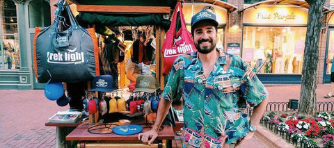 A man in a floral shirt and Trek Light cap outside a Trek Light market booth, packed hammocks hanging behind him