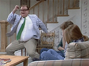 A man in a plaid jacket and tie dancing energetically in a living room, with two people seated nearby.