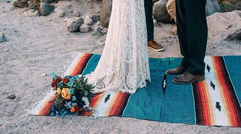 A bride and groom standing on a teal striped Trek Light blanket in the desert, with a bouquet resting beside them