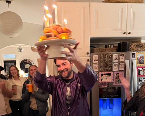 A person holding a cake with lit candles at a celebration, people clapping and smiling in the kitchen.