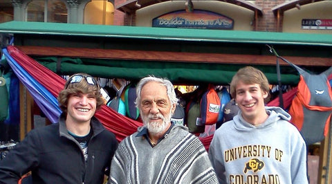 Tommy Chong in a striped poncho standing between two smiling young men in front of the Trek Light hammock kiosk in Boulder