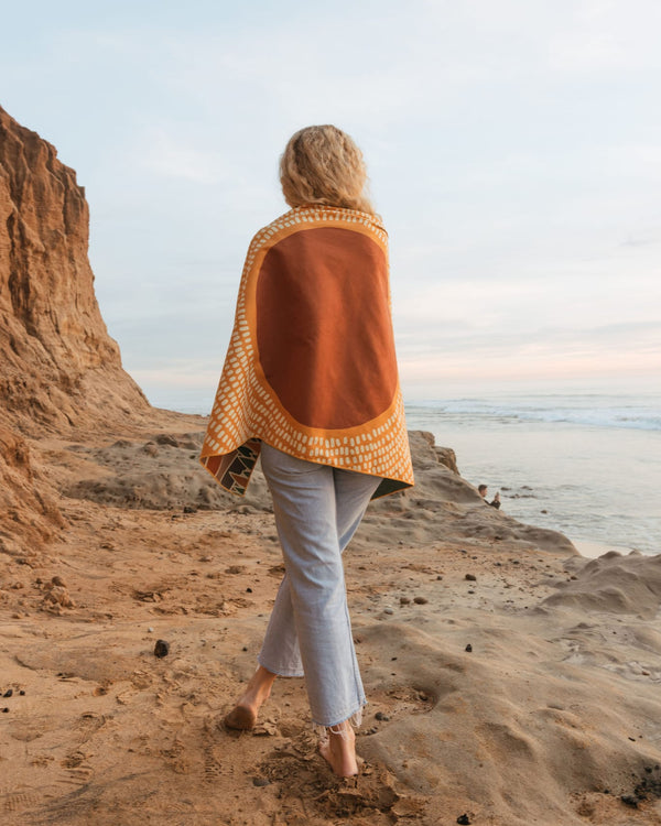 Woman wrapped in Trek Light blanket standing by the seaside cliffs during sunset.