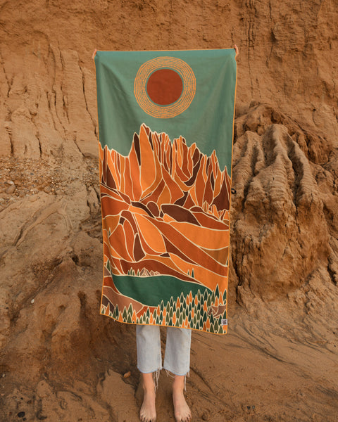 Trek Light towel with mountain and sun design held in front of rocky backdrop, blending nature and art for outdoor adventures.
