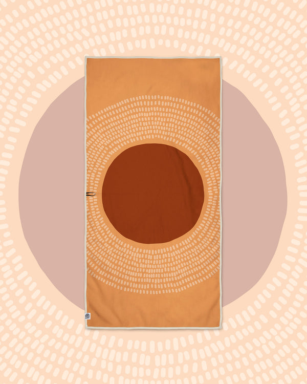 Alpenglow Wander & Travel Towel in orange with a large rust circle and dotted rays, on a peach and mauve sun print backdrop