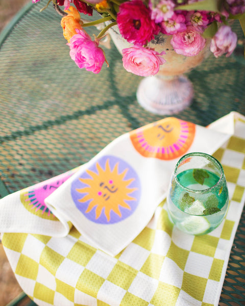 Always Sunny Kitchen & Tea Towel displayed on a table with a floral arrangement and a drink.