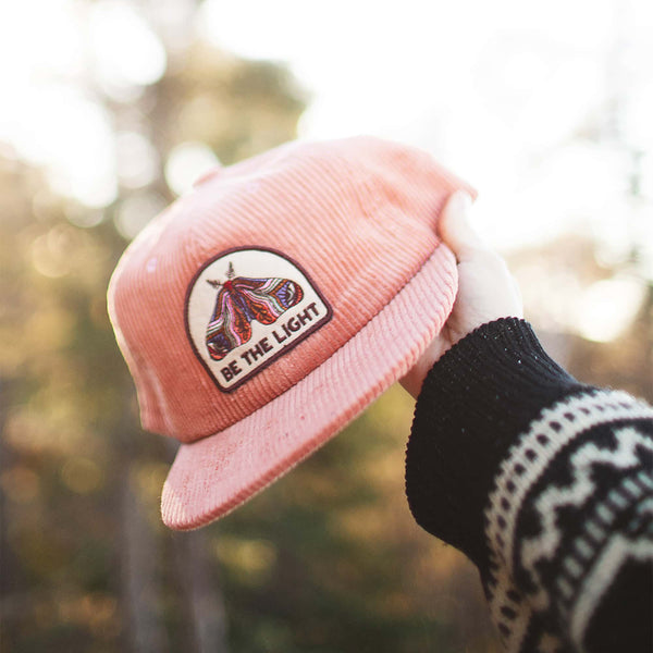 Person holding a coral Trek Light 6-panel snapback hat with a "Be The Light" moth design, flat brim, perfect for outdoor adventures.