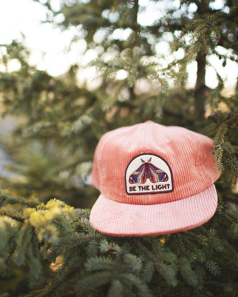 Be The Light Hat resting on a tree branch, showcasing its corduroy design and vivid embroidery.