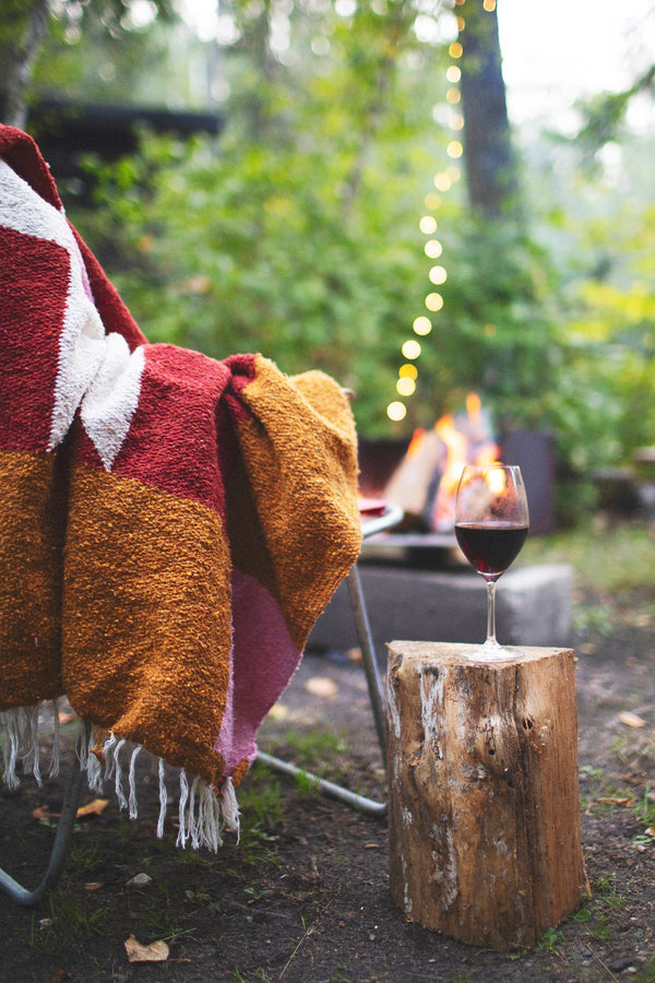 Cozy Bernie Blanket draped over chair by campfire, glass of wine on log. Handwoven, made from recycled materials; perfect for relaxing. By Trek Light.