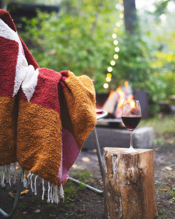 Cozy Bernie Blanket draped over chair by campfire, glass of wine on log. Handwoven, made from recycled materials; perfect for relaxing. By Trek Light.