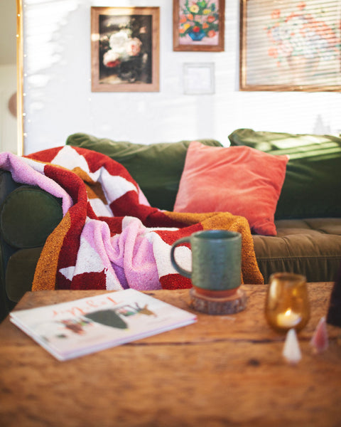 Cozy living room setup featuring the Bernie Blanket draped on a green couch with decorative pillows and a coffee mug.