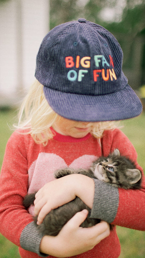 Child wearing a hat that says 'BIG FAN OF FUN' while holding a kitten.