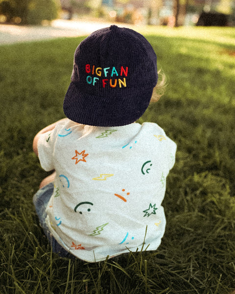 Child wearing a Big Fan Of Fun Kids Hat sitting in grass, showcasing colorful embroidery and playful design.