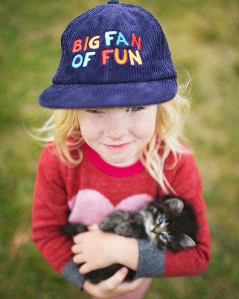 Child wearing a Big Fan Of Fun Kids Hat while holding a kitten in a park setting.