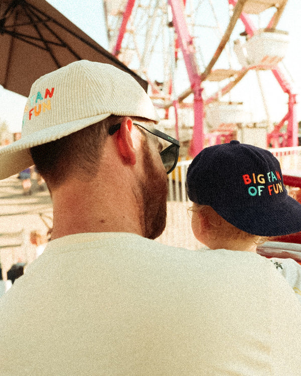 Big Fan Of Fun Hat Twinsie Set, cream and navy corduroy caps with colorful letters, worn by an adult and child at a fair.