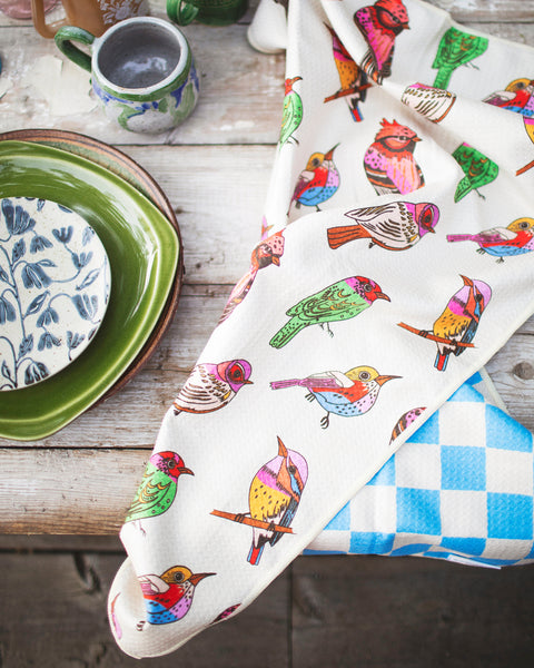 Bird Song Kitchen & Tea Towel showcasing vibrant bird designs on a kitchen table with plates and mugs.