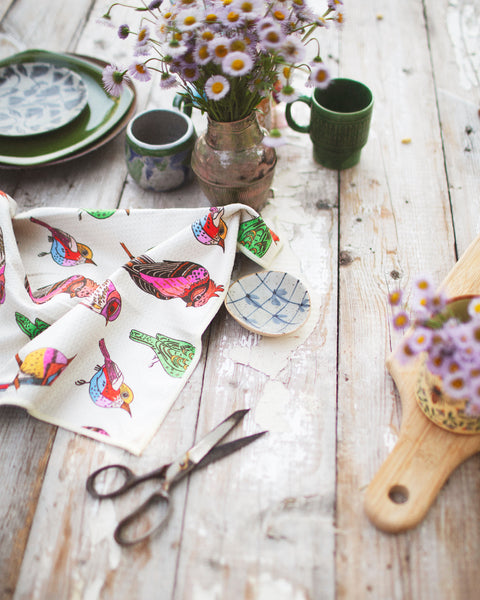 Bird Song Kitchen & Tea Towel displayed with plates, cups, scissors, and flowers on a wooden table.