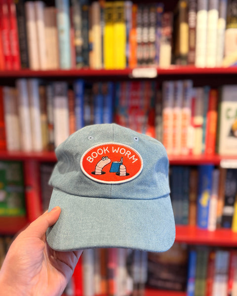 Person holding a Book Worm Hat with an illustration of worms reading in front of a colorful bookshelf.