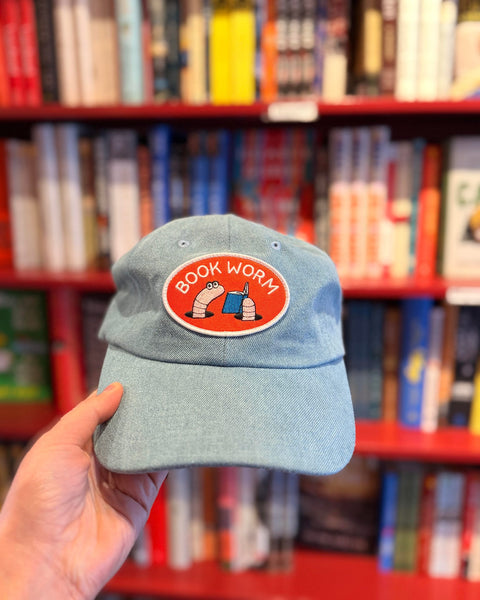 Person holding a Book Worm Hat with an illustration of worms reading in front of a colorful bookshelf.