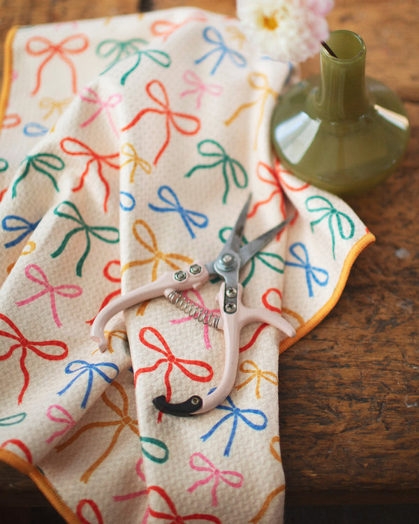 Holiday Bows Kitchen & Tea Towel with colorful bow designs, scissors, and a green vase on a wooden table.