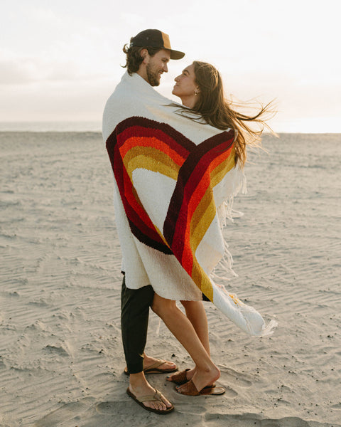 Couple wrapped in Trek Light's Bowie Blanket with vintage rainbow design, enjoying a beach moment.