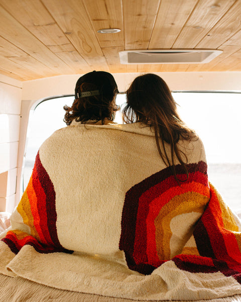Couple wrapped in a Trek Light Bowie Blanket with rainbow design, enjoying a cozy moment in a wooden van interior.