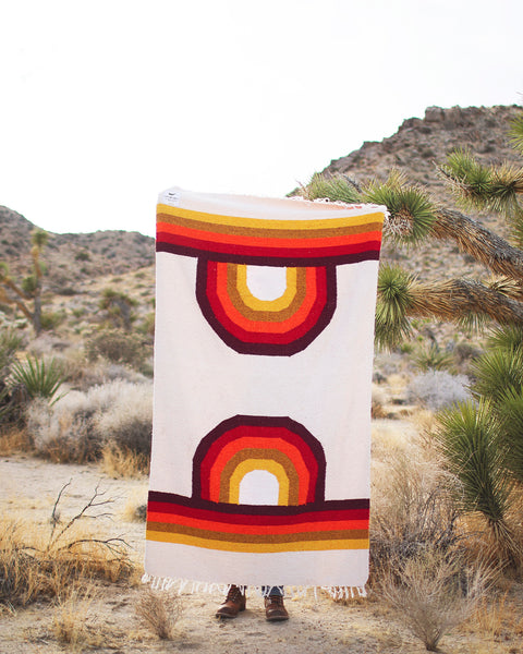 A person holding a Bowie Blanket with vibrant rainbow design in a desert landscape.