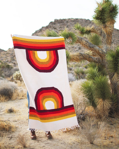 Person holding a Bowie Blanket outdoors, showcasing vibrant rainbow design in a desert landscape.