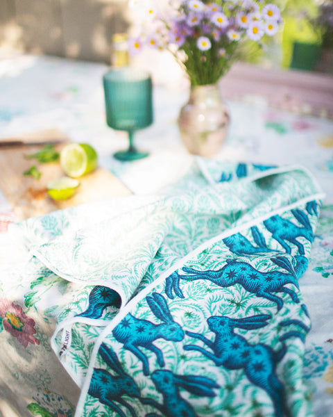 Bunnies On Parade Kitchen & Tea Towel displayed on a table with flowers and a drink, showcasing its vibrant design.