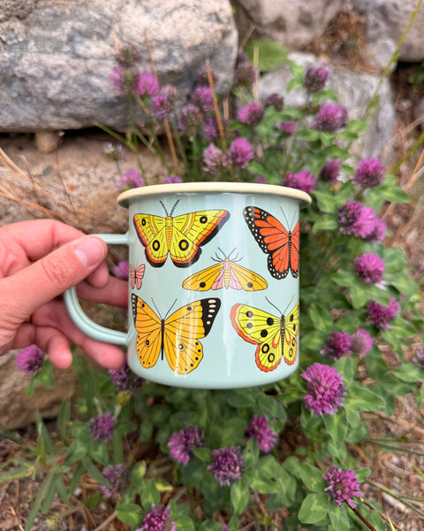 Butterfly Beauty Mug - Enamel 16oz Mug held in hand surrounded by purple flowers and rocks.