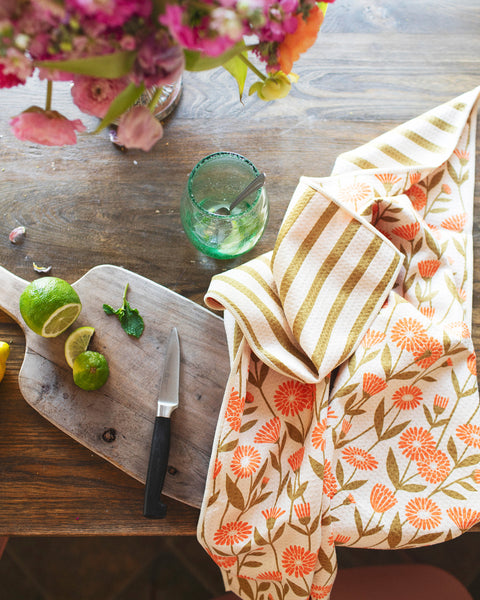 Calendula Flowers Kitchen & Tea Towel displayed on a wooden table with limes and a floral arrangement.