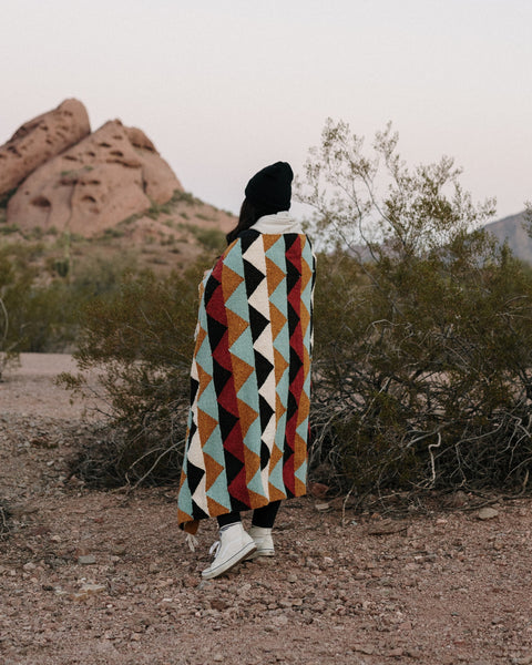 Person wrapped in a Cascade Blanket with teal, rust, red, black and white triangles, in a desert with rocks.