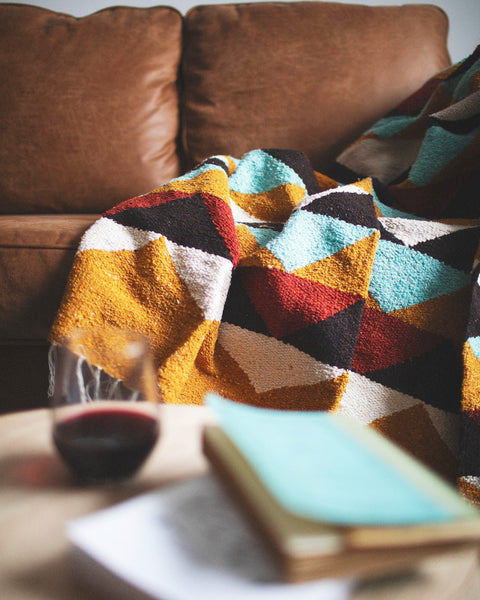 Beautiful handwoven Cascade Blanket made from recycled materials, featuring a colorful, cozy design on a sofa with a book and wine glass nearby. By Trek Light.