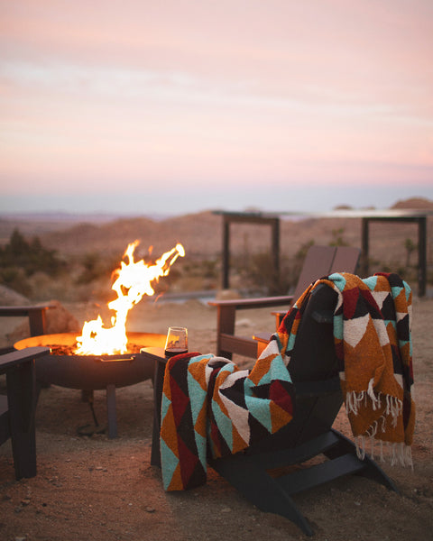 A colorful geometric patterned handwoven throw blanket is draped over a dark wooden chair next to a lit fire pit in an outdoor desert setting at sunset.