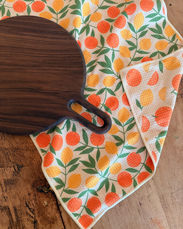 Saved By Citrus Kitchen & Tea Towel with vibrant citrus design and wooden cutting board on a rustic wooden table.
