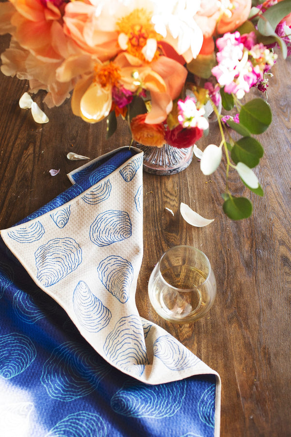 Coastal Oysters Kitchen & Tea Towel displayed with a floral arrangement and glass on a wooden table.