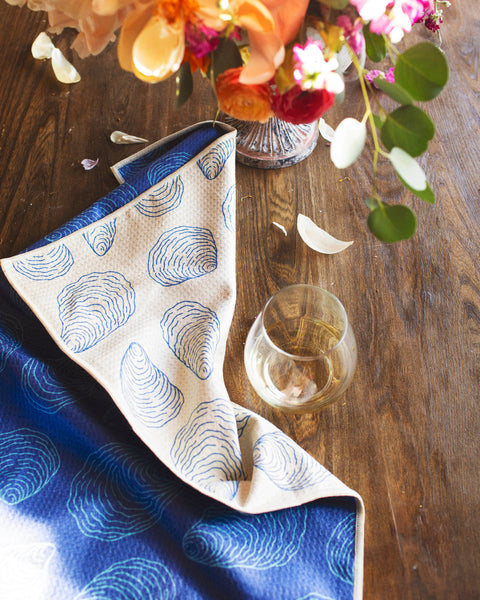 Coastal Oysters Kitchen & Tea Towel displayed with a floral arrangement and glass on a wooden table.