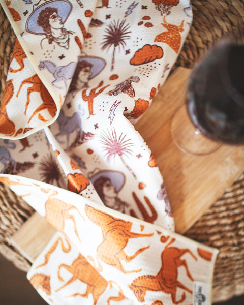 Cosmic Cowgirl Kitchen & Tea Towel with vibrant designs and eco-friendly material on a wooden table.