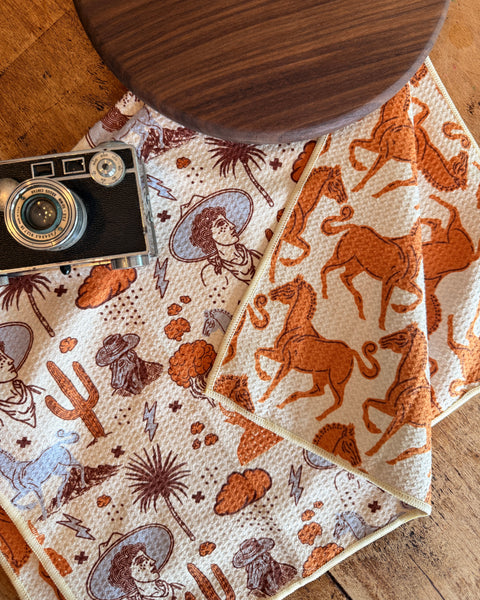 Cosmic Cowgirl Kitchen & Tea Towel with vibrant western designs and super absorbent fabric on a wooden table.