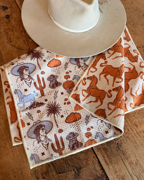 Cosmic Cowgirl Kitchen & Tea Towel featuring vibrant western designs and a stylish hat on a wooden table.