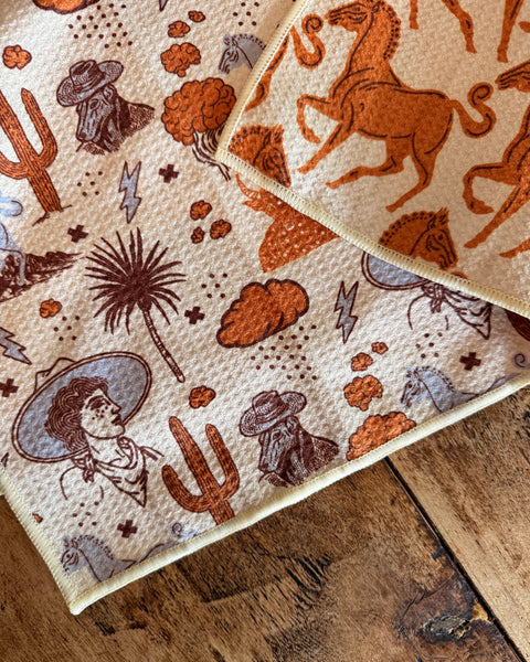 Cosmic Cowgirl Kitchen & Tea Towel featuring vibrant western designs with horses, cowboys, and cacti on a textured fabric.