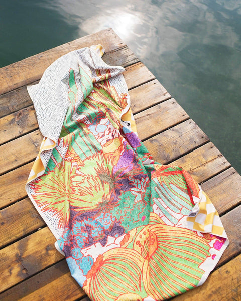 Desert Dreams travel towel with a colorful cactus print and yellow checked edges lying on a wooden dock beside lake water.