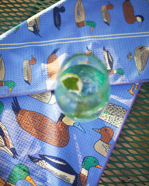 Ducks In A Row Kitchen & Tea Towel with colorful duck designs and a glass of drink on a table.