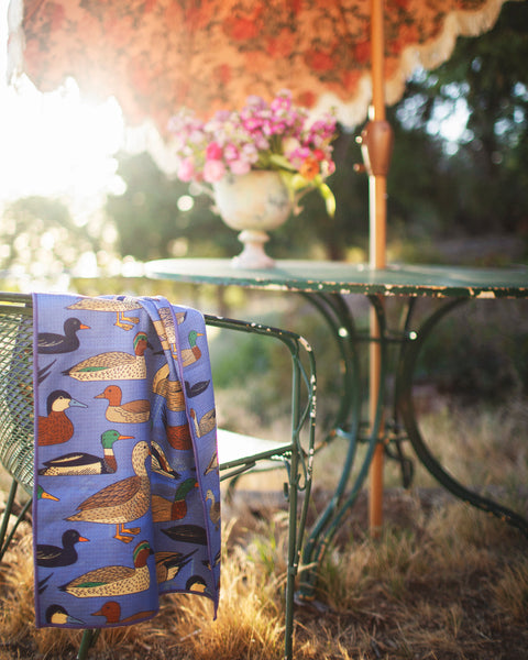 Ducks In A Row Kitchen & Tea Towel draped over a chair next to a table with flowers, showcasing vibrant duck designs.