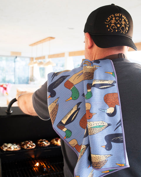 Person using Ducks In A Row Kitchen & Tea Towel while grilling, showcasing vibrant duck designs and practicality.