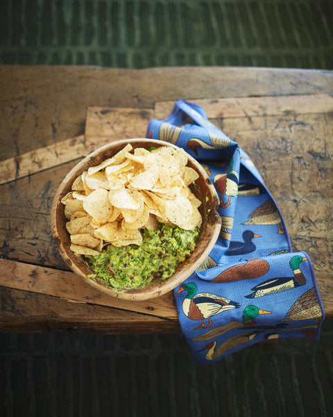 Ducks In A Row Kitchen & Tea Towel next to a bowl of guacamole and tortilla chips on a wooden table.