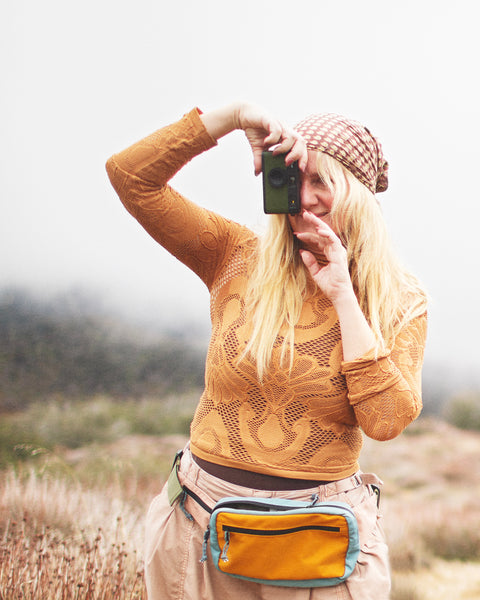 Woman taking a photo outdoors wearing an Everyday Fanny Pack (Golden Hour) in a scenic background.
