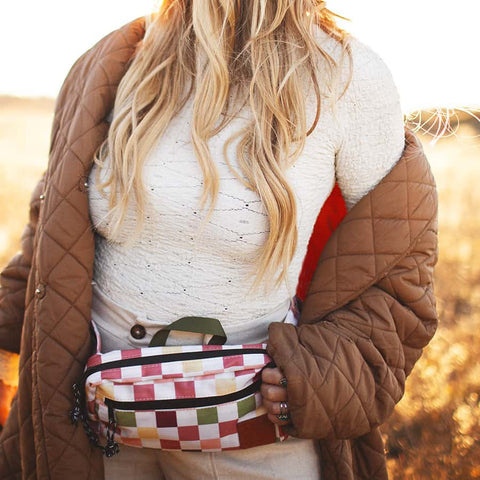 Woman wearing a quilted brown jacket and holding a checkered bag while standing in a sunlit field.
