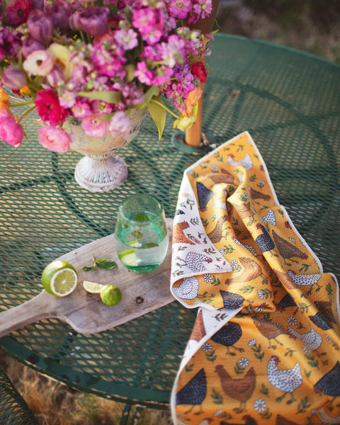 Farmhouse Chicken Kitchen & Tea Towel displayed on a green table with flowers and a drink.