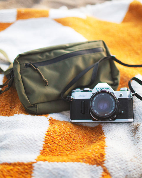 Everyday Fanny Pack (Fern) next to a Canon camera on a patterned blanket at the beach.